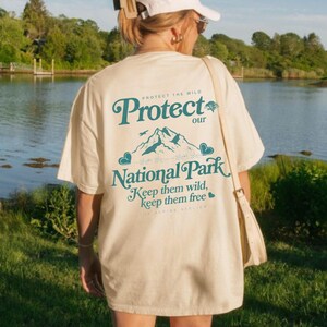 May include: Cream-colored t-shirt with a teal graphic that reads "Protect our National Park." The design includes mountains, birds, and hearts. The text "Keep them wild, keep them free" is also featured. The Alpine Atelier is printed at the bottom.