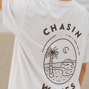 以下が含まれることがあります： 背面にダークブラウンのグラフィックがプリントされた白い半袖Tシャツ。楕円形のフレームの中にヤシの木、三日月、波が描かれています。楕円の上部には「CHASIN」、下部には「WAVES」という文字が配置されています。
