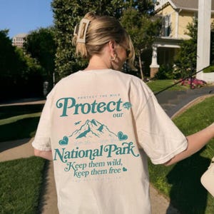 May include: Cream-colored t-shirt with teal text and graphics. The text reads "Protect our National Park" with mountain and heart illustrations. The shirt also has the phrases "Keep them wild, keep them free" and "Protect the Wild".