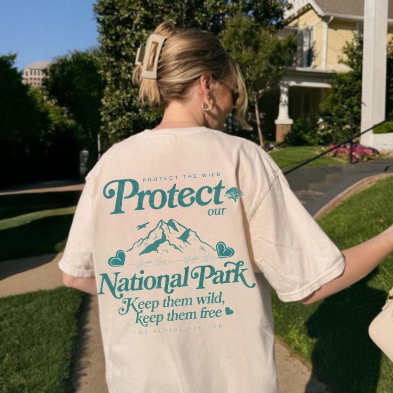 May include: Cream-colored t-shirt with teal text and graphics. The text reads "Protect our National Park" with mountain and heart illustrations. The shirt also has the phrases "Keep them wild, keep them free" and "Protect the Wild".