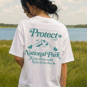 May include: White t-shirt with a teal graphic on the back that reads "Protect our National Park." The design includes mountain illustrations, hearts, and the text "Keep them wild, keep them free."