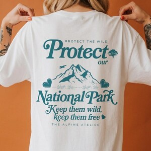 May include: White t-shirt with teal text and graphics. The design includes the words "Protect our National Park" with a mountain illustration, a tree, and the phrase "Keep them wild, keep them free." The Alpine Atelier is also printed on the shirt.