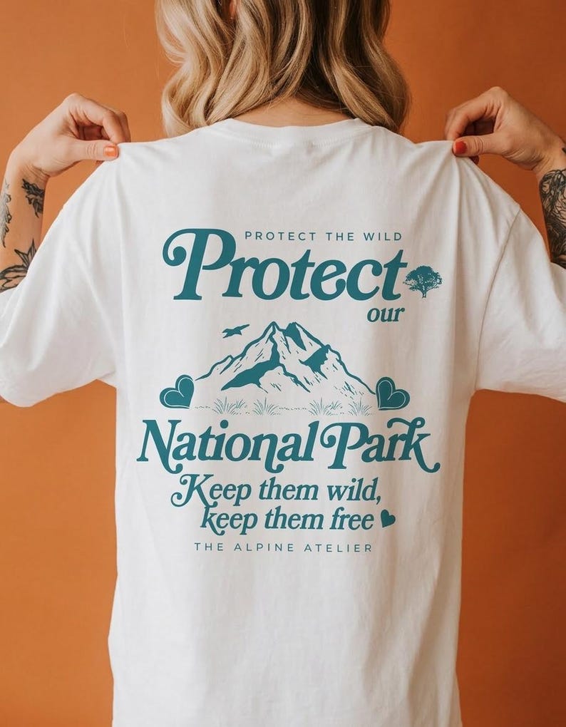 May include: White t-shirt with teal text and graphics. The design includes the words "Protect our National Park" with a mountain illustration, a tree, and the phrase "Keep them wild, keep them free." The Alpine Atelier is also printed on the shirt.