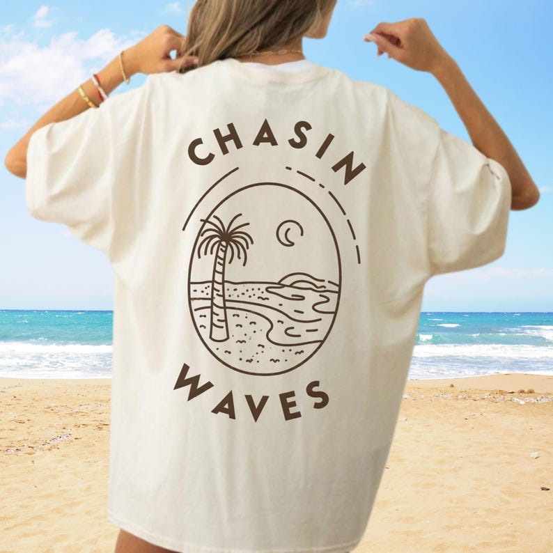 以下が含まれることがあります： オフホワイトの半袖Tシャツで、背面にダークブラウンのグラフィックがプリントされています。ヤシの木、三日月、波が描かれた円形のフレームと、太字のサンセリフ体で「CHASIN WAVES」というテキストがデザインされています。