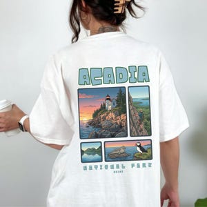 May include: White t-shirt featuring the word "ACADIA" in light blue block letters above four rectangular images. The images depict scenes from Acadia National Park, including a lighthouse, mountain views, and wildlife. The words "NATIONAL PARK" and "MAINE" are also visible.