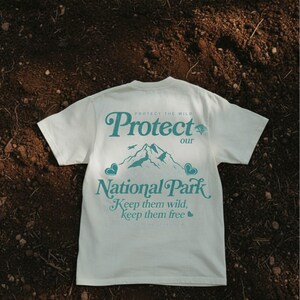 May include: Cream-colored t-shirt with the text "Protect our National Park" in teal. The design includes a mountain range and the phrases "Keep them wild, keep them free". The shirt is short-sleeved.