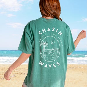 以下が含まれることがあります： 背面に白いグラフィックがプリントされた緑色の半袖Tシャツ。円形のフレームの中にヤシの木、三日月、波が描かれており、上下に「CHASIN WAVES」という文字がアーチ状に配置されています。