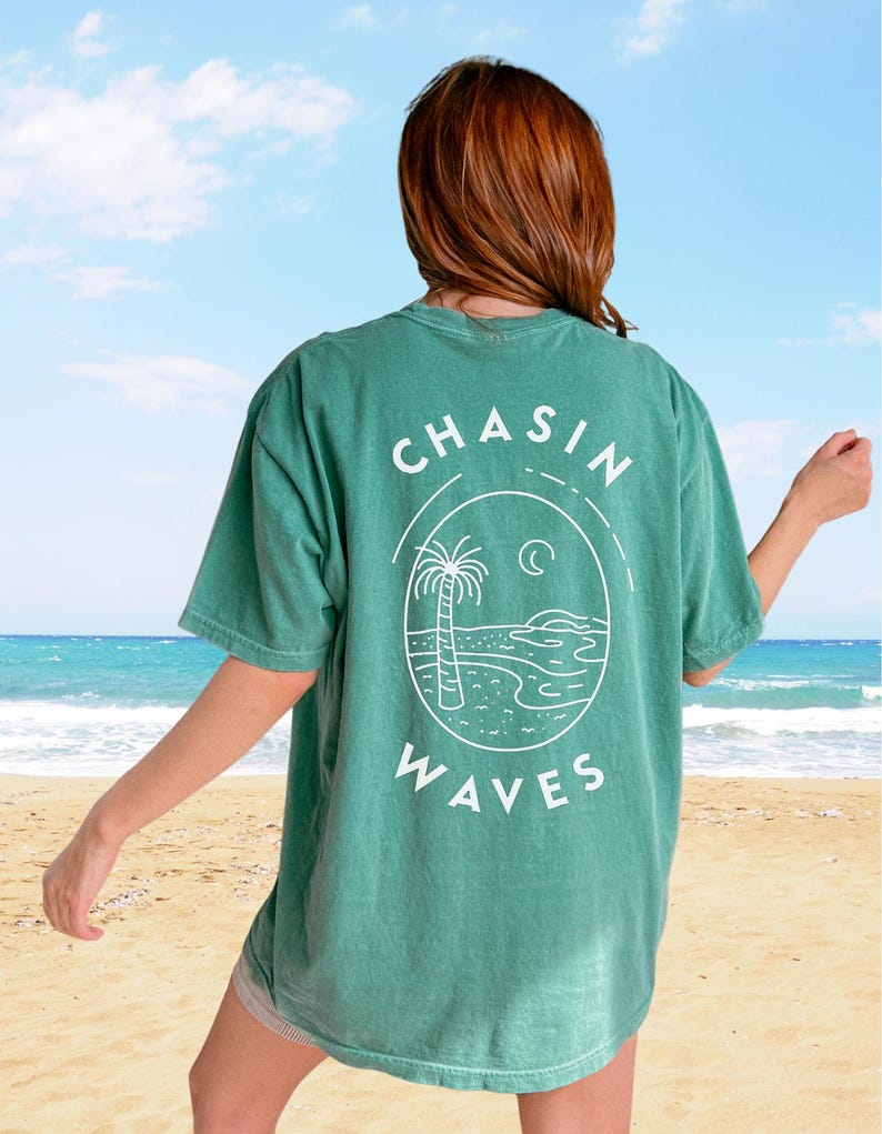 以下が含まれることがあります： 背面に白いグラフィックがプリントされた緑色の半袖Tシャツ。円形のフレームの中にヤシの木、三日月、波が描かれており、上下に「CHASIN WAVES」という文字がアーチ状に配置されています。