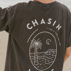 以下が含まれることがあります： 背面に白いグラフィックが施されたチャコールグレーの半袖Tシャツ。デザインには「CHASIN WAVES」の文字がアーチ状に描かれ、ヤシの木、三日月、様式化された海岸線が描かれた楕円形のフレームを囲んでいます。