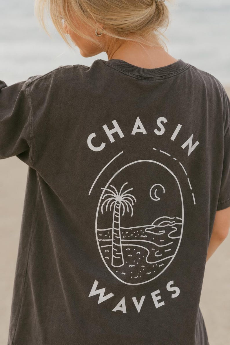 以下が含まれることがあります： 背面に白いグラフィックが施されたチャコールグレーの半袖Tシャツ。デザインには「CHASIN WAVES」の文字がアーチ状に描かれ、ヤシの木、三日月、様式化された海岸線が描かれた楕円形のフレームを囲んでいます。