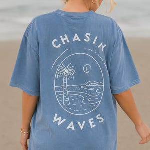 以下が含まれることがあります： 背面に白いラインアートのグラフィックが施された青い半袖Tシャツ。デザインには、ヤシの木、三日月、そして円形のイラストの周りに太字でアーチ状に配置された「CHASIN WAVES」というテキストが描かれたビーチの風景が含まれています。