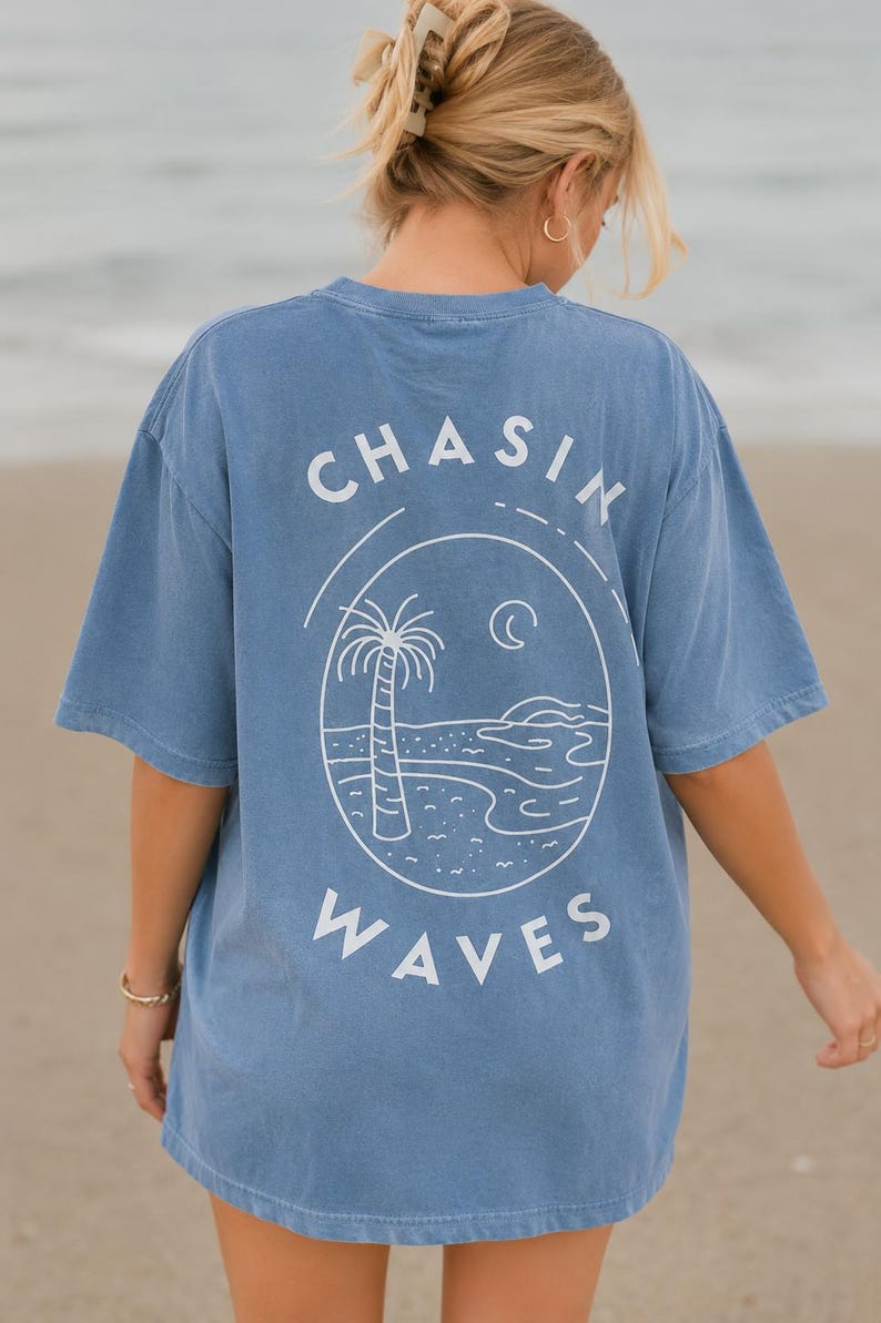 以下が含まれることがあります： 背面に白いラインアートのグラフィックが施された青い半袖Tシャツ。デザインには、ヤシの木、三日月、そして円形のイラストの周りに太字でアーチ状に配置された「CHASIN WAVES」というテキストが描かれたビーチの風景が含まれています。