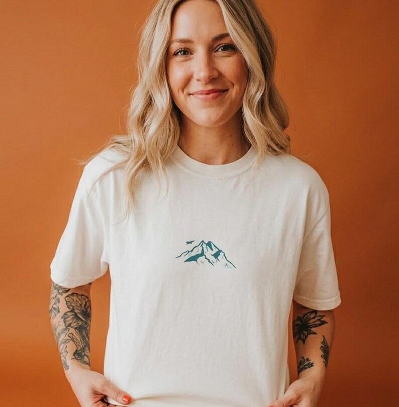 May include: Cream-colored t-shirt featuring a teal mountain graphic with a bird. The shirt has a crew neck and short sleeves. The design is centered on the chest. The model is smiling.