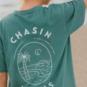以下が含まれることがあります： 背面に白いグラフィックが施されたティールカラーの半袖Tシャツ。デザインには「CHASIN WAVES」という文字が含まれており、ヤシの木、三日月、波打ち際のあるビーチの風景が描かれた楕円形のフレームを囲んでいます。