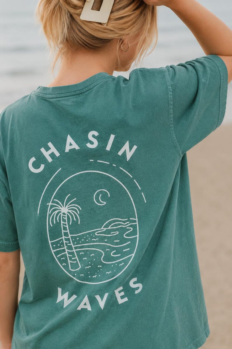以下が含まれることがあります： 背面に白いグラフィックが施されたティールカラーの半袖Tシャツ。デザインには「CHASIN WAVES」という文字が含まれており、ヤシの木、三日月、波打ち際のあるビーチの風景が描かれた楕円形のフレームを囲んでいます。