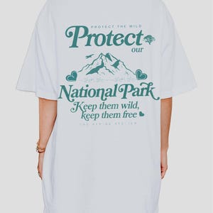 May include: White t-shirt with the text "Protect our National Park" in teal. The design includes a mountain range, a bird, and hearts. The text "Keep them wild, keep them free" is also featured. The Alpine Atelier is printed at the bottom.