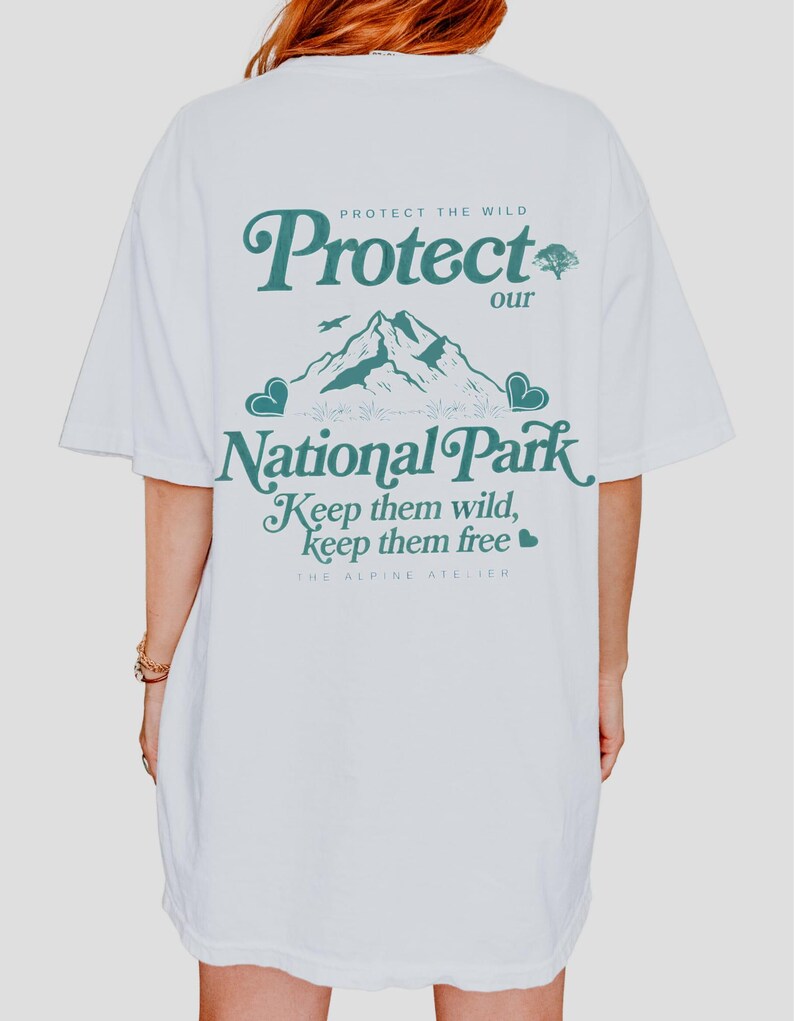 May include: White t-shirt with the text "Protect our National Park" in teal. The design includes a mountain range, a bird, and hearts. The text "Keep them wild, keep them free" is also featured. The Alpine Atelier is printed at the bottom.