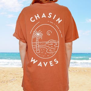 以下が含まれることがあります： 背面に白いラインアートのグラフィックが施された、錆色の半袖Tシャツ。デザインは楕円形のフレームの中にヤシの木、三日月、海の波が描かれており、白い大文字で「CHASIN WAVES」というテキストがプリントされています。