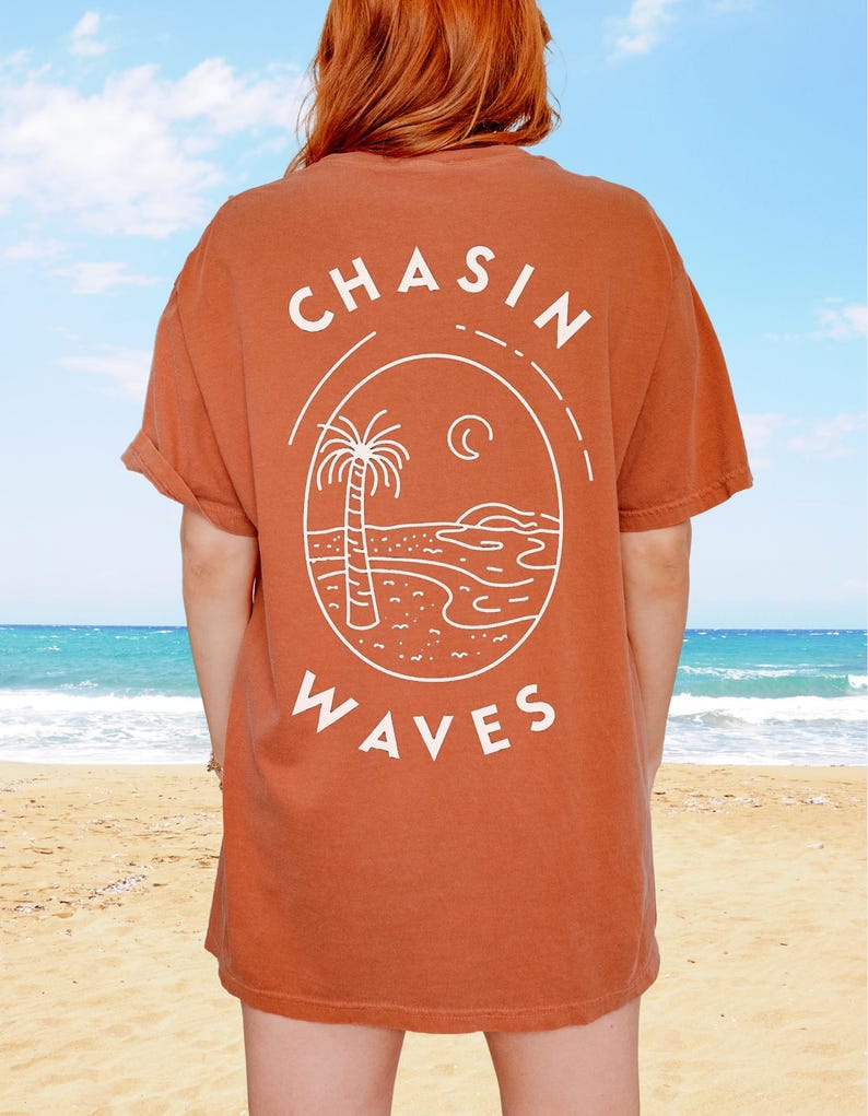 以下が含まれることがあります： 背面に白いラインアートのグラフィックが施された、錆色の半袖Tシャツ。デザインは楕円形のフレームの中にヤシの木、三日月、海の波が描かれており、白い大文字で「CHASIN WAVES」というテキストがプリントされています。