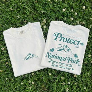 May include: Two white t-shirts with green mountain graphics and text. One shirt has a small mountain graphic, the other says "Protect our National Park" with mountains, hearts, and the text "Keep them wild, keep them free."