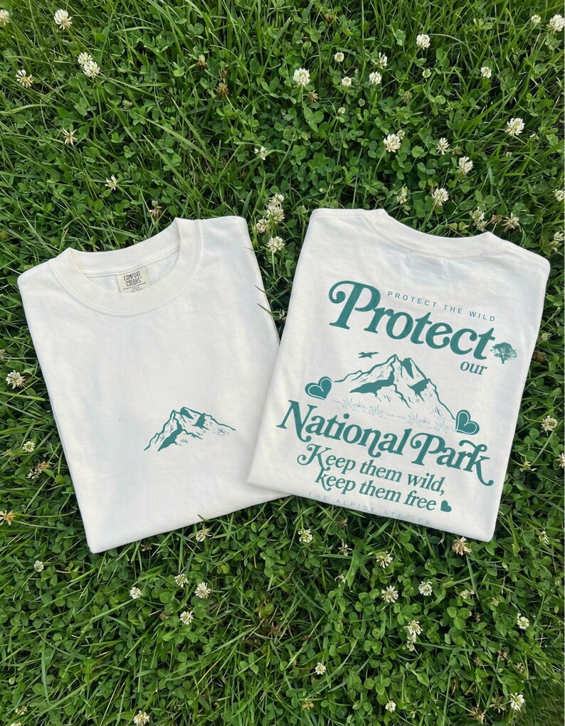 May include: Two white t-shirts with green mountain graphics and text. One shirt has a small mountain graphic, the other says "Protect our National Park" with mountains, hearts, and the text "Keep them wild, keep them free."