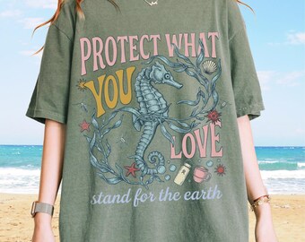 以下が含まれることがあります： オリーブグリーンのTシャツ。タツノオトシゴ、海藻、貝殻、ヒトデ、水筒、マグカップのグラフィックプリントが施されています。テキストはピンク、黄色、白の文字で「PROTECT WHAT YOU LOVE」および「stand for the earth」と書かれています。