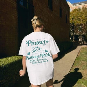 May include: White t-shirt with the text "Protect our National Park" in green. The design includes mountains and the phrase "Keep them wild, keep them free." The shirt is a casual, oversized fit.