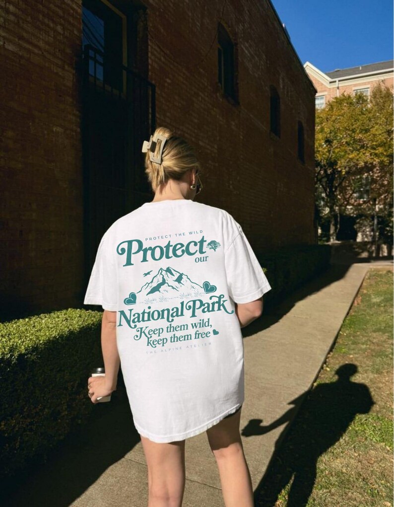 May include: White t-shirt with the text "Protect our National Park" in green. The design includes mountains and the phrase "Keep them wild, keep them free." The shirt is a casual, oversized fit.