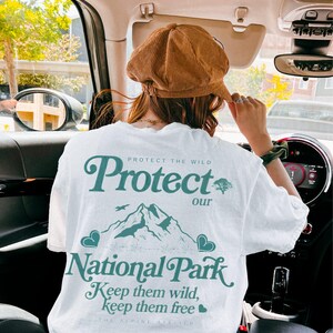 May include: White t-shirt with a teal graphic that reads "Protect our National Park." The design includes a mountain range, birds, and hearts. The shirt also has the text "Keep them wild, keep them free."