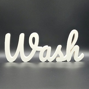 Può includere: Cartello in legno color crema con la parola "Wash" in un carattere corsivo. Il cartello è esposto su uno sfondo grigio scuro. Le lettere sono collegate e hanno un aspetto liscio e arrotondato, adatto all'arredamento del bagno.