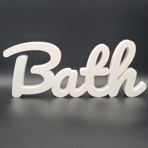 May include: A white wooden sign in a cursive font, spelling the word "Bath". The sign is set against a dark background, with a smooth, clean finish on the letters. Ideal for bathroom decoration, this sign adds a touch of elegance.