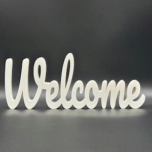 以下が含まれることがあります： クリーム色の筆記体で書かれた「Welcome」のサイン。単語は流れるような筆記体で書かれており、文字はつながっています。サインはしっかりとした素材でできており、暗い背景に配置されています。