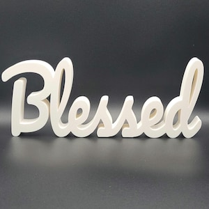 Puede incluir: Letrero de madera color crema con la palabra "Blessed" escrita en cursiva. El letrero es independiente y tiene un acabado liso y mate. Las letras están conectadas, creando un diseño fluido y elegante. El fondo es negro.
