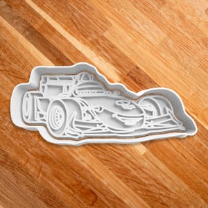 May include: A white plastic cookie cutter shaped like a race car. The cookie cutter is detailed with a driver's helmet and a spoiler.