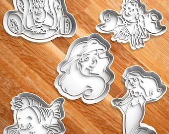 May include: Five white plastic cookie cutters in various shapes. The shapes include Ursula, Flounder, Sebastian, and two versions of Ariel from The Little Mermaid. The cutters are on a wooden surface.