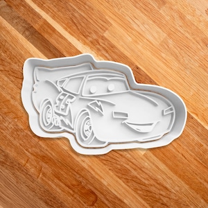 May include: White plastic cookie cutter shaped like a cartoon race car with a lightning bolt design. The car has a smiling face with two eyes and a wide mouth.