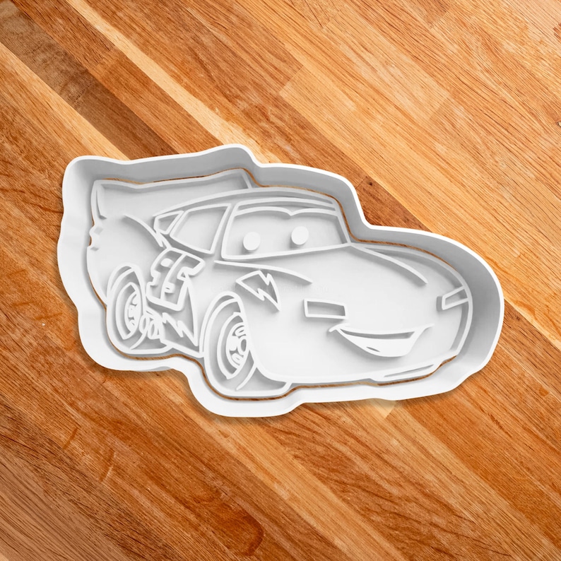 May include: White plastic cookie cutter shaped like a cartoon race car with a lightning bolt design. The car has a smiling face with two eyes and a wide mouth.