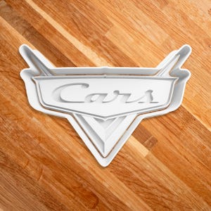 May include: White plastic cookie cutter shaped like the Cars logo with the word "Cars" in a stylized font.
