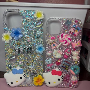 Puede incluir: Dos fundas transparentes para teléfono decoradas con pedrería, purpurina y abalorios coloridos. Una funda presenta un diseño de Hello Kitty con un lazo azul, mientras que la otra presenta un diseño de Hello Kitty con un lazo rosa. Ambas fundas tienen la palabra "ERIN" escrita en pedrería plateada.