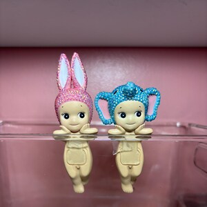 Puede incluir: Dos peque&ntilde;as figuras, una con orejas de conejo rosas y otra con orejas de elefante azules, ambas adornadas con adornos brillantes. Las figuras tienen caras sonrientes y son de color beige claro. Se exhiben sobre una superficie transparente.