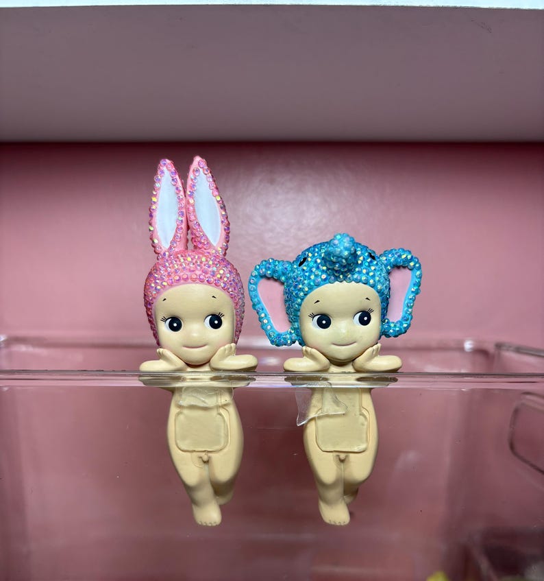 Puede incluir: Dos peque&ntilde;as figuras, una con orejas de conejo rosas y otra con orejas de elefante azules, ambas adornadas con adornos brillantes. Las figuras tienen caras sonrientes y son de color beige claro. Se exhiben sobre una superficie transparente.