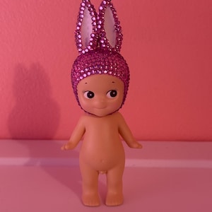 Puede incluir: Peque&ntilde;a figura desnuda con una cara sonriente y ojos grandes. La figura lleva un tocado de orejas de conejo rosa enjoyado. El fondo es de color rosa liso.