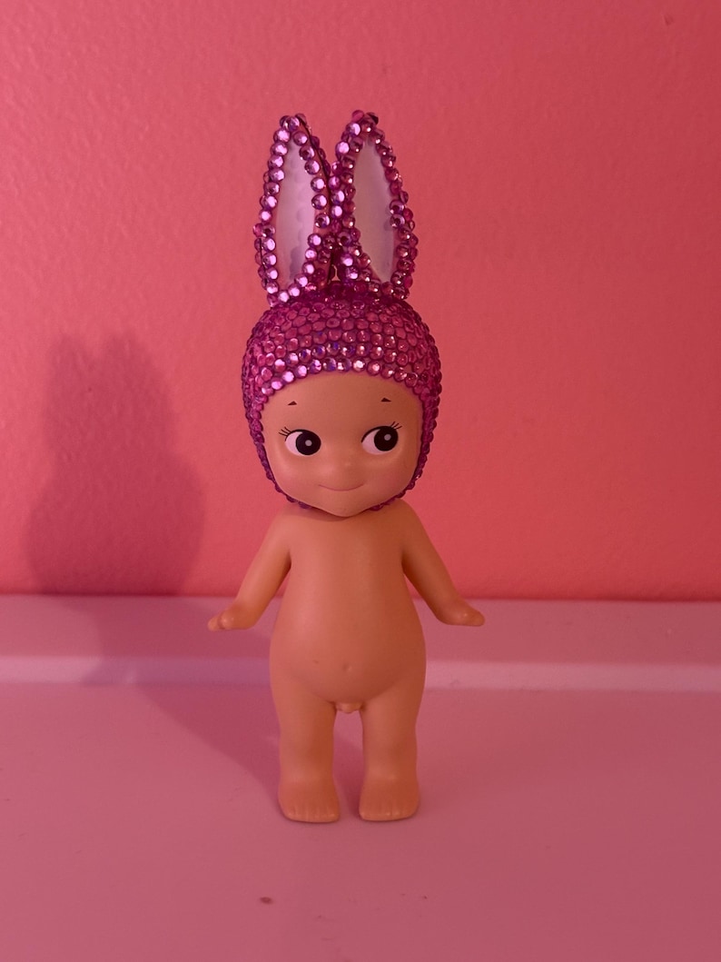 Puede incluir: Peque&ntilde;a figura desnuda con una cara sonriente y ojos grandes. La figura lleva un tocado de orejas de conejo rosa enjoyado. El fondo es de color rosa liso.