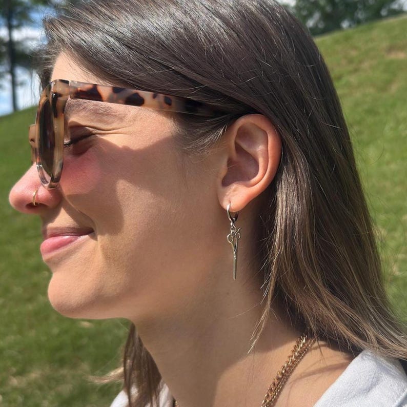 May include: A pair of silver earrings with a scissor design hanging from a hoop. The earrings are worn by a person with brown hair and a tortoiseshell patterned pair of sunglasses.