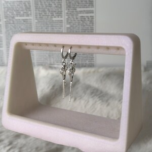May include: A pair of silver hoop earrings with a key charm dangling from each hoop. The earrings are displayed on a white earring stand.