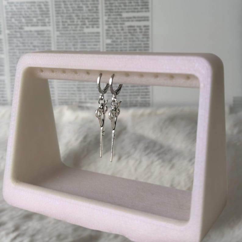 May include: A pair of silver hoop earrings with a key charm dangling from each hoop. The earrings are displayed on a white earring stand.