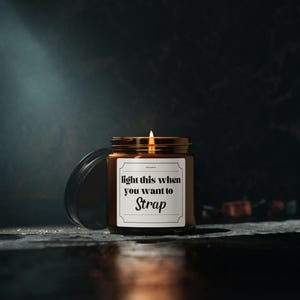 Puede incluir: Una vela de vidrio marrón encendida con tapa dorada y etiqueta blanca. La etiqueta dice "light this when you want to Strap". La vela está sobre una superficie oscura con un fondo oscuro.