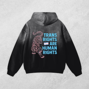 May include: Black hoodie with a gradient effect. The back features a pink and black tiger graphic and the text "TRANS RIGHTS ARE HUMAN RIGHTS" in blue, with a transgender flag. The hoodie is a statement piece.