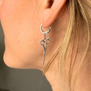 May include: Silver hoop earrings with a dangling pair of silver scissors charm.