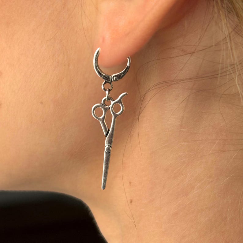 May include: Silver hoop earrings with a pair of scissors dangling from the bottom.